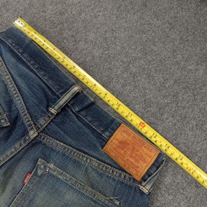 以下が含まれることがあります： 黄色いメジャーが付いた青いデニムジーンズのクローズアップ。ジーンズには「JOE MCCOY & CO.」の文字と寸法が書かれた茶色の革パッチが付いています。テープは約38cmの測定値を示しています。