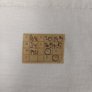May include: A close-up of a white fabric with a small, rectangular, tan tag in the lower center. The tag has handwritten numbers and letters in black ink, including "168", "34", and "190". There are also circles with letters inside.