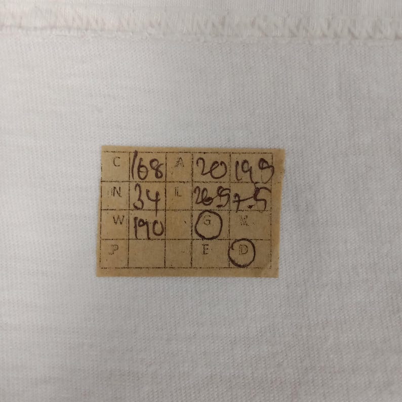 May include: A close-up of a white fabric with a small, rectangular, tan tag in the lower center. The tag has handwritten numbers and letters in black ink, including "168", "34", and "190". There are also circles with letters inside.