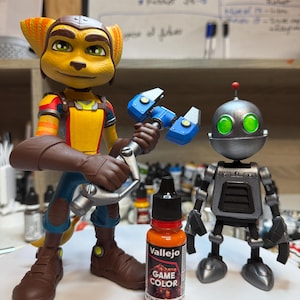 May include: A detailed image featuring two character figurines: a yellow and brown anthropomorphic creature holding a blue tool, and a silver robot with green eyes. A bottle of orange Vallejo Game Color paint and a paintbrush are in the foreground.