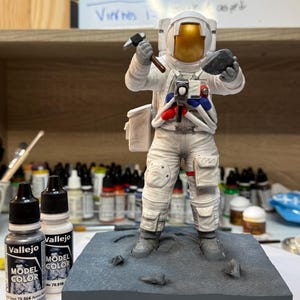 May include: A detailed model of an astronaut in a white spacesuit, holding a hammer and a rock, standing on a gray base. The base is inscribed with "APOLLO 17". Two bottles of Vallejo Model Color paint are in the foreground.
