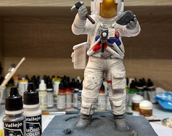 May include: A detailed model of an astronaut in a white spacesuit, holding a hammer and a rock, standing on a gray base. The base is inscribed with "APOLLO 17". Two bottles of Vallejo Model Color paint are in the foreground.
