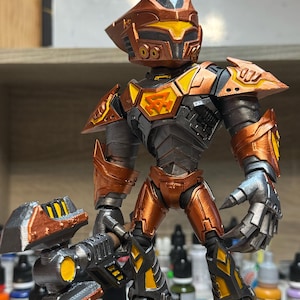 Op de afbeelding: Gedetailleerd actiefiguur van een robotstrijder, met een metallic kleurenschema van koper, zilver en gele accenten. Het figuur poseert met een groot wapen en ingewikkelde pantserdetails. De helm van de robot heeft een geometrisch ontwerp.