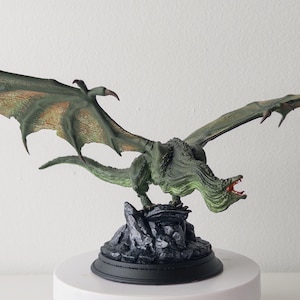 May include: A detailed figurine of a dragon with outstretched wings. The dragon is primarily green with hints of orange and red, perched atop a rocky base. The wings are spread wide, showcasing intricate details.