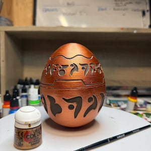 May include: A copper-colored, egg-shaped container with carved, stylized symbols. The container is sitting on a white surface next to a small paint bottle and a thin paintbrush. The paint bottle has the text "CE COLOUR" and "12+".