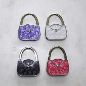 May include: Four handbag holders shaped like purses, in purple, white, black, and red. Each has a silver handle and a swirl pattern. A small gem is set in the centre of each purse.