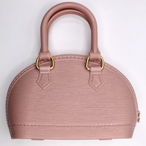 Könnte beinhalten: Eine staubrosa Handtasche mit einer strukturierten, abgerundeten Form. Sie hat zwei obere Griffe und goldfarbene Beschläge. Die Tasche hat eine strukturierte Oberfläche und einen Reißverschluss.