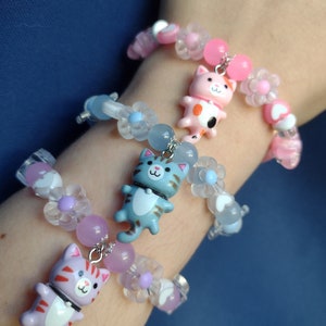May include: Three bracelets with cat charms. The bracelets are made of clear beads with pink, blue, and purple accents. The cat charms are pink, blue, and purple.