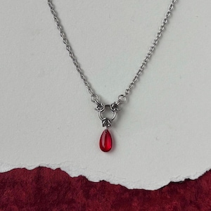 Pode incluir: Um colar prateado com um pingente vermelho em forma de gota. O pingente pende de um grupo de três anéis. A corrente é delicada e o fundo é papel branco texturizado sobre uma superfície vermelha.