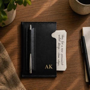 May include: Black leather wallet with a pen holder and a notepad. The wallet has the initials "AK" in gold lettering. A silver pen is in the pen holder. The notepad has the text "Idea for a new minimalist wallet design - clean edges."