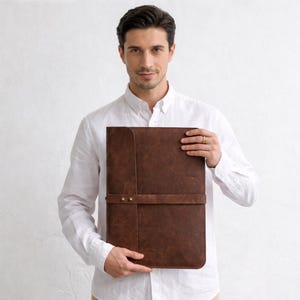 May include: A brown leather portfolio with a strap closure, held by a person wearing a white button-down shirt. The portfolio has a textured surface and a vertical seam down the front.