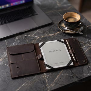 May include: A dark brown leather folio case holding a digital notepad with the text "SUPER NOTE". A silver pen is held in place. A laptop and a coffee cup are visible in the background.