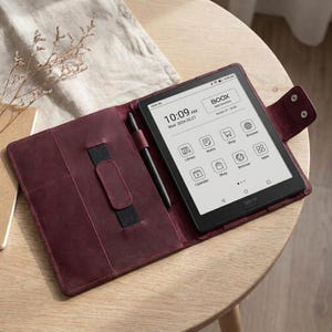 May include: A burgundy leather e-reader case with a black e-reader and stylus. The e-reader displays the time, date, and app icons. The case has a strap to hold the stylus and a snap closure. The brand name "BOOX" is visible.