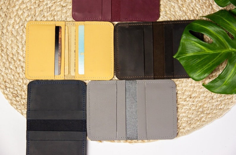May include: Six different leather card holders in various colors, including yellow, brown, gray, black, and burgundy. The card holders are laid out on a woven straw surface.