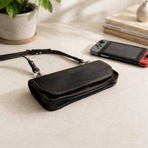 Op de afbeelding: Een donkerbruine leren hoes met witte stiksels en een bijpassende riem. De hoes is ontworpen om een Nintendo Switch-console te bevatten, die op de achtergrond zichtbaar is. De rechthoekige hoes heeft een gebogen ontwerp.