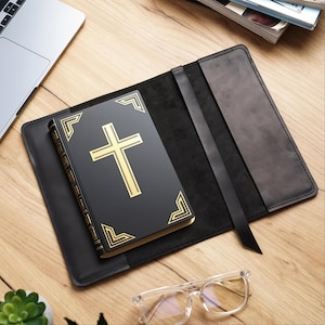 Puede incluir: Un libro negro con una cruz dorada y detalles decorativos en las esquinas, colocado en un estuche de cuero negro abierto con una cinta negra como marcapáginas. El libro está sobre una superficie de madera, con un portátil y gafas en el fondo.