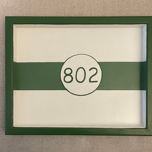 May include: A green wooden frame with a white background and a green stripe. The number 802 is written in a circle in the center of the frame.