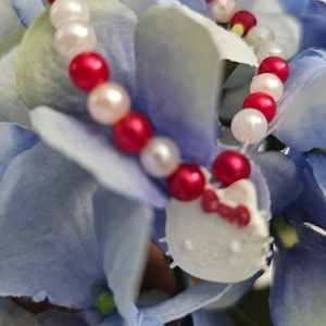 Könnte beinhalten: Ein rotes und weißes Perlenarmband mit einem Hello-Kitty-Anhänger. Das Armband liegt auf einer blauen und weißen Blume.