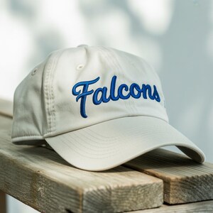 May include: A cream-colored baseball cap with the word "Falcons" embroidered in blue script across the front. The cap has a curved brim and a classic six-panel design. The hat is resting on a wooden surface.