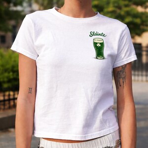 K&ouml;nnte beinhalten: Wei&szlig;es T-Shirt mit einer gr&uuml;nen Abbildung eines Bierkrugs und einem Kleeblatt. Das Wort "Sl&aacute;inte" ist &uuml;ber dem Bierglas aufgedruckt. Das Shirt ist kurz&auml;rmlig und aus weichem Material.