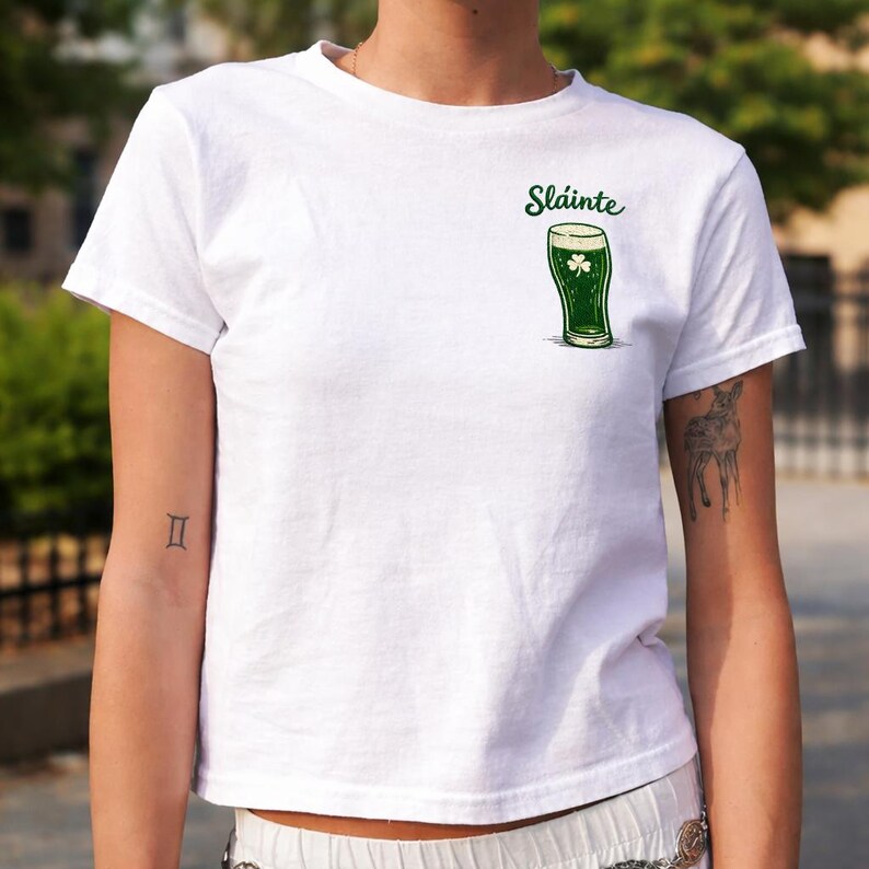 K&ouml;nnte beinhalten: Wei&szlig;es T-Shirt mit einer gr&uuml;nen Abbildung eines Bierkrugs und einem Kleeblatt. Das Wort "Sl&aacute;inte" ist &uuml;ber dem Bierglas aufgedruckt. Das Shirt ist kurz&auml;rmlig und aus weichem Material.