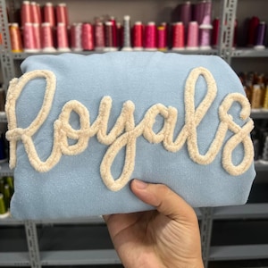 May include: A light blue sweatshirt with the word "Royals" embroidered in white fluffy yarn.