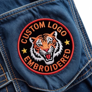 May include: Embroidered patch featuring a tiger design with the text "CUSTOM LOGO EMBROIDERED". The patch is round with a black background, orange tiger, and red border. The patch is sewn onto a blue denim fabric.