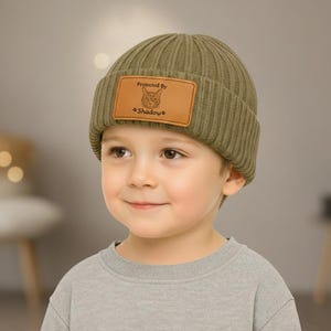 May include: A ribbed olive green beanie with a brown leather patch. The patch features a cat illustration and the text "Protected By Shadows". The beanie is worn by a young child.