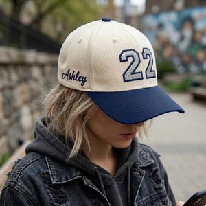 May include: A cream-colored baseball cap with a dark blue brim. The front features the number 22 in a silver glitter finish, and the side is embroidered with the name Ashley in dark blue script lettering. The cap has a structured crown.