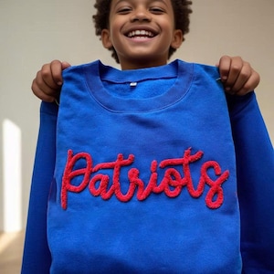 May include: A royal blue sweatshirt with the word "Patriots" in red, stylized script. The sweatshirt has a crew neck and long sleeves. The image shows the sweatshirt being held up.