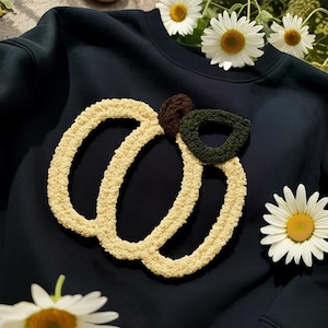 May include: Navy blue sweatshirt with a cream-colored, crocheted pumpkin design. The pumpkin has a brown stem and a green leaf. White daisies are scattered around the sweatshirt, creating a fall theme.