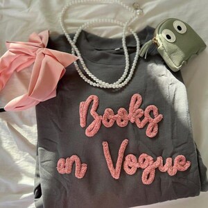 May include: A gray sweatshirt with the text "Books in Vogue" in pink, paired with a pink bow headband, a pearl necklace, and a small green coin purse with eyes.