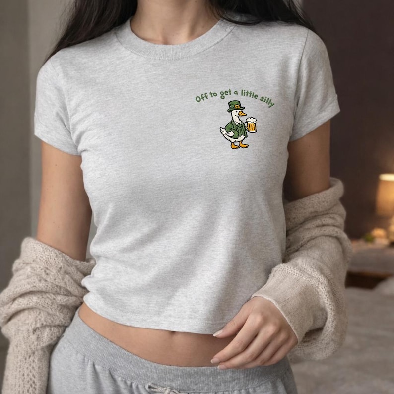 K&ouml;nnte beinhalten: Hellgraues T-Shirt mit einer Cartoon-Ente in gr&uuml;ner Jacke und Hut, die einen Bierkrug h&auml;lt. Der Text "Off to get a little silly" steht &uuml;ber der Ente. Das T-Shirt hat kurze &Auml;rmel.