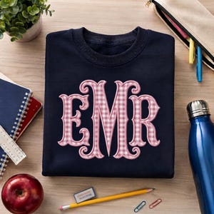 May include: A navy blue crewneck sweatshirt featuring a large, centered monogram with the letters EMR. The letters are rendered in a decorative, serif font with a light pink and white gingham checkered pattern and a solid pink outline.