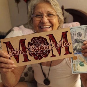 May include: Wooden sign with the word "MOM" and a rose design, with the names "John", "Sophia", and "Alex". The sign is held with a $100 bill.