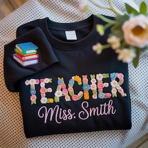 May include: Black sweatshirt with floral embroidered "TEACHER" and "Miss. Smith" in pink script. A stack of books is embroidered on the sleeve. The sweatshirt is a cozy and stylish gift for teachers.