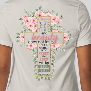 Puede incluir: Camiseta de color claro con un diseño de cruz adornada con rosas rosas y hojas verdes. La cruz contiene el texto "Engañosa es la gracia, y vana la hermosura; la mujer que teme a Jehová, ésa será alabada" de Proverbios 31:30.