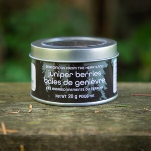 May include: A silver tin with a black label that reads "SEASONINGS FROM THE HEARTLAND juniper berries baies de genièvre LES ASSAISONNEMENTS DU TERROIR Net WT 20 g POIDS net".