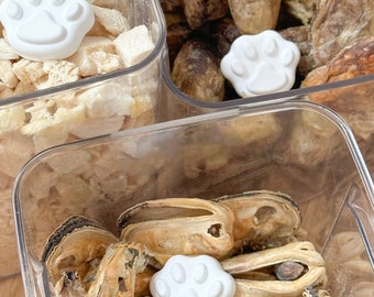May include: Three clear plastic containers filled with different types of dog treats. The containers have paw print lids. The first container has white chicken pieces, the second container has brown dried meat, and the third container has dried mussel shells.