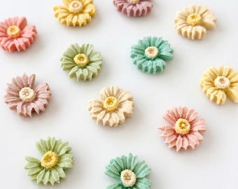 May include: A collection of colorful, flower-shaped decorations in various pastel shades. The flowers have a textured appearance with a yellow center and petals in pink, yellow, green, and peach. The arrangement is on a white background.