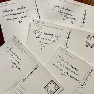 Könnte beinhalten: Eine Sammlung weißer Postkarten mit handgeschriebenen Nachrichten und dekorativen Elementen. Die Nachrichten enthalten Sätze wie "Es gibt einen göttlichen Plan der Güte" und "Vertraue dem Prozess des Lebens". Die Postkarten sind auf einer dunklen Holzoberfläche angeordnet.