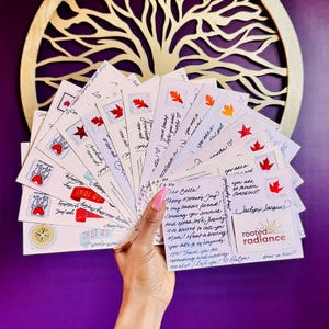 May include: A hand holds a fan of postcards with handwritten messages and maple leaf stamps. The postcards are white with red and orange accents. A gold tree of life decorates the background.