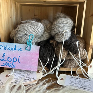 Könnte beinhalten: Eine Holzkiste gefüllt mit Knäueln isländischer Lopi-Wolle in verschiedenen Grautönen und Brauntönen. Die Wolle ist mit "Icelandic Lopi Yarn" beschriftet.
