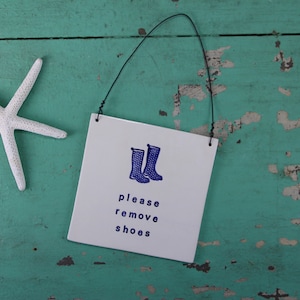 May include: A white starfish and a white square sign with a black wire hanger. The sign has a blue illustration of rain boots and the text "please remove shoes" in blue. The background is a distressed, turquoise-colored wooden surface.