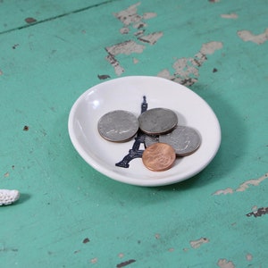 May include: A white ceramic dish with a black Eiffel Tower design holds four coins. The dish is on a teal blue wooden surface.