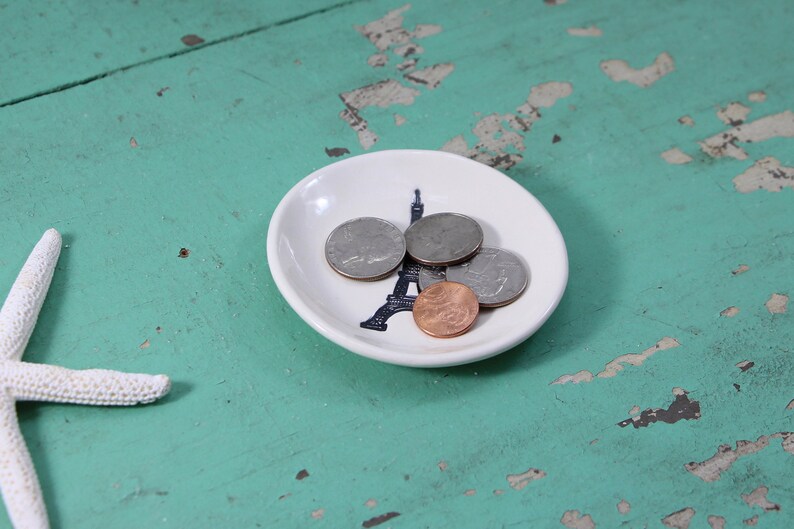 May include: A white ceramic dish with a black Eiffel Tower design holds four coins. The dish is on a teal blue wooden surface.