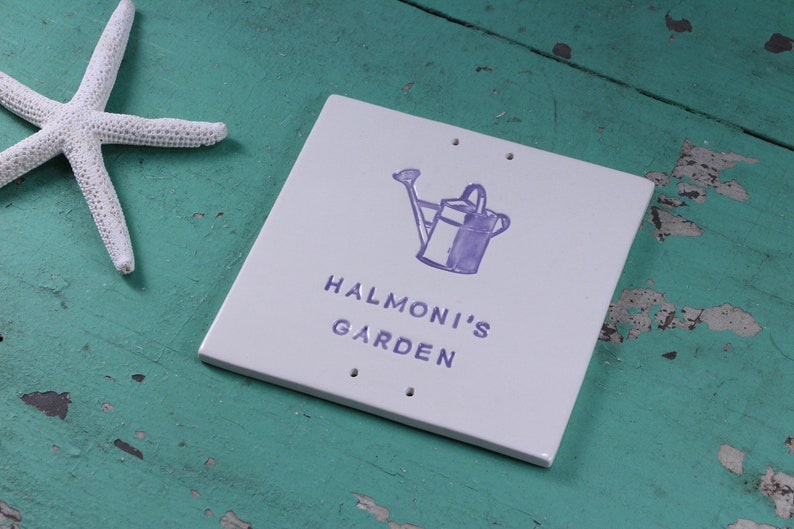 May include: White ceramic garden sign with purple lettering that reads "Halmoni's Garden". A purple watering can with a flower is above the text.