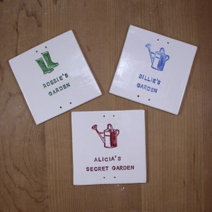 May include: Three white ceramic garden signs with green, blue, and red lettering. The signs feature a pair of green rain boots, a blue watering can, and a red watering can. The signs read "Robbie's Garden", "Billie's Garden", and "Alicia's Secret Garden".