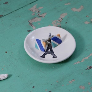 May include: A white ceramic dish with a black and white Eiffel Tower design. Two cufflinks are on the dish. The cufflinks are triangular and have blue, orange, and white stripes.