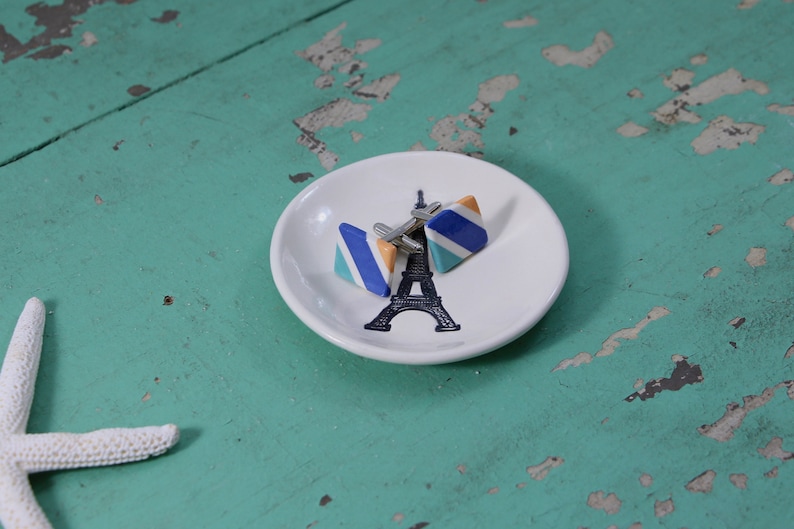 May include: A white ceramic dish with a black and white Eiffel Tower design. Two cufflinks are on the dish. The cufflinks are triangular and have blue, orange, and white stripes.
